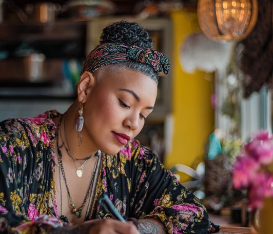 Woman writing in a journal at a warm, sunlit table with colorful clothing and natural decor