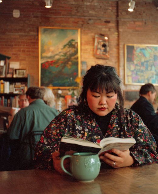 Woman reading a book in a warm café with a green ceramic mug, brick walls, and art in background